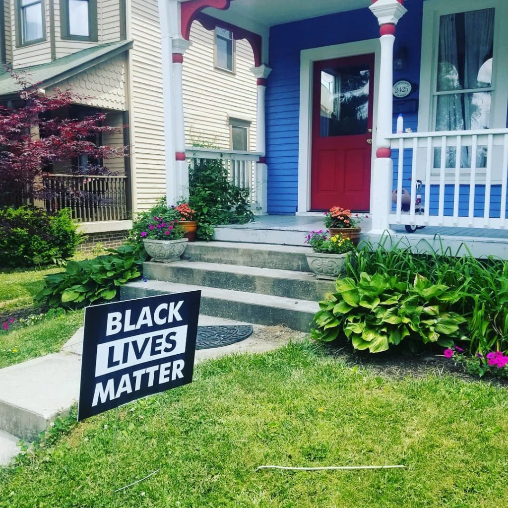 the front yard of a brightly-painted house with a verdant garden, sloppy grass, and a Black Lives Matter sign, on a sunny day